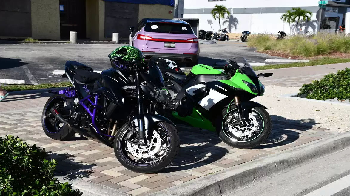 Two sport motorcycles, one black and one green, parked on a sidewalk in an urban area with a car in the background.