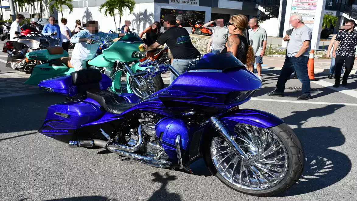 Vibrant blue custom motorcycle at a gathering with several people in the background on a sunny day.