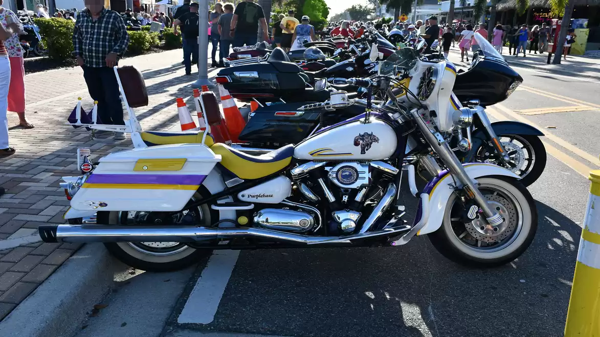 A white motorcycle with yellow and purple accents is parked on a street next to other motorcycles. People and trees are visible in the background.