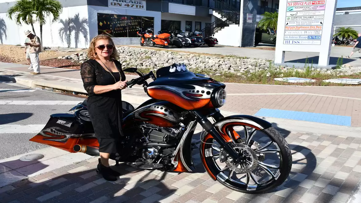 A woman in black stands next to a striking custom motorcycle with orange and black flame designs, on a paved walkway.