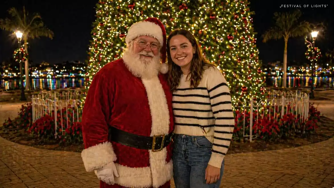Man in Santa Claus costume stands next to a woman in front of a large, illuminated Christmas tree on a festively decorated square.