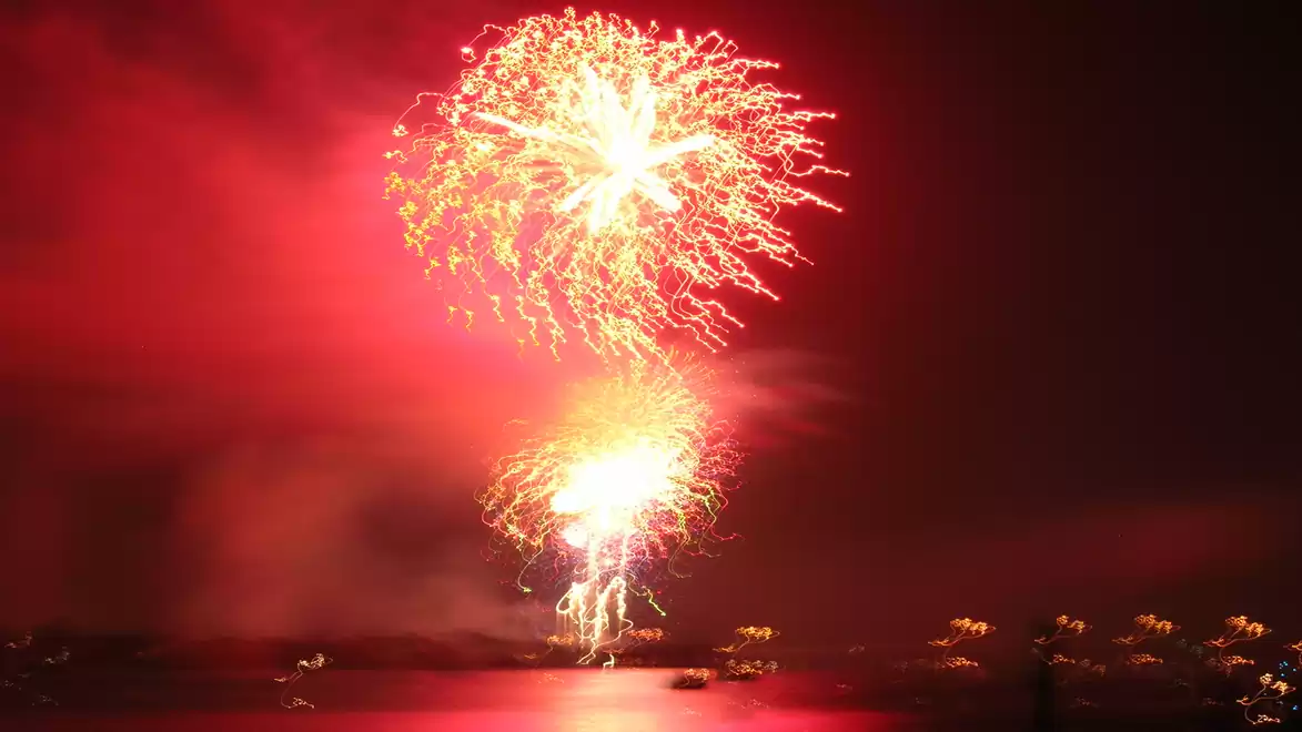 A colorful fireworks display lights up the night sky above a calm body of water, reflecting the lights.