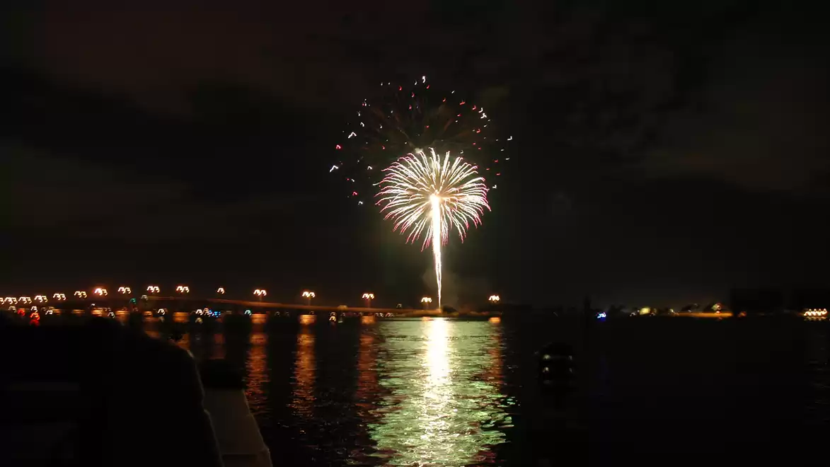 A firework illuminates the night sky over a lit bridge and reflects on the water.