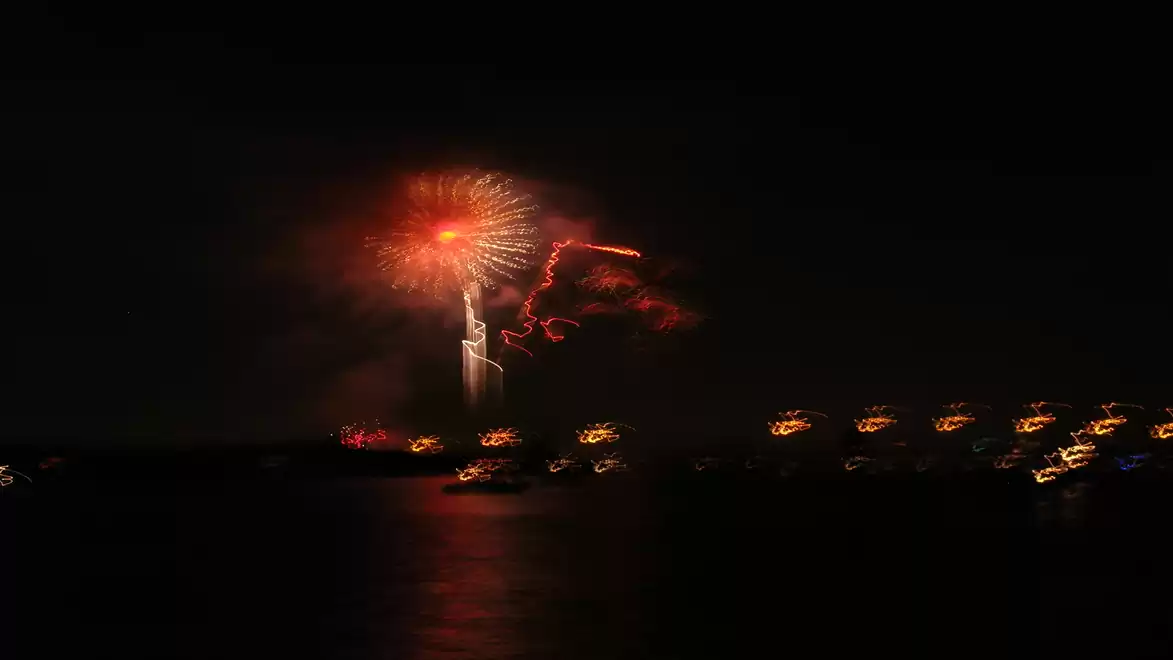 A fireworks display illuminates the night sky over a body of water with illuminated structures in the background.
