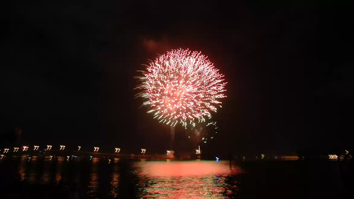 A large firework illuminates the night sky over a body of water, with lights reflecting on the shore.