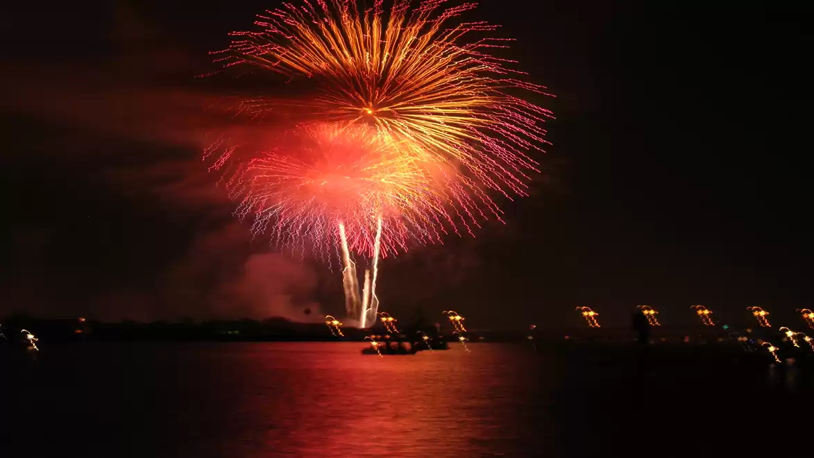 A vibrant firework display over a dark body of water at night with reflections of lights on the water surface.