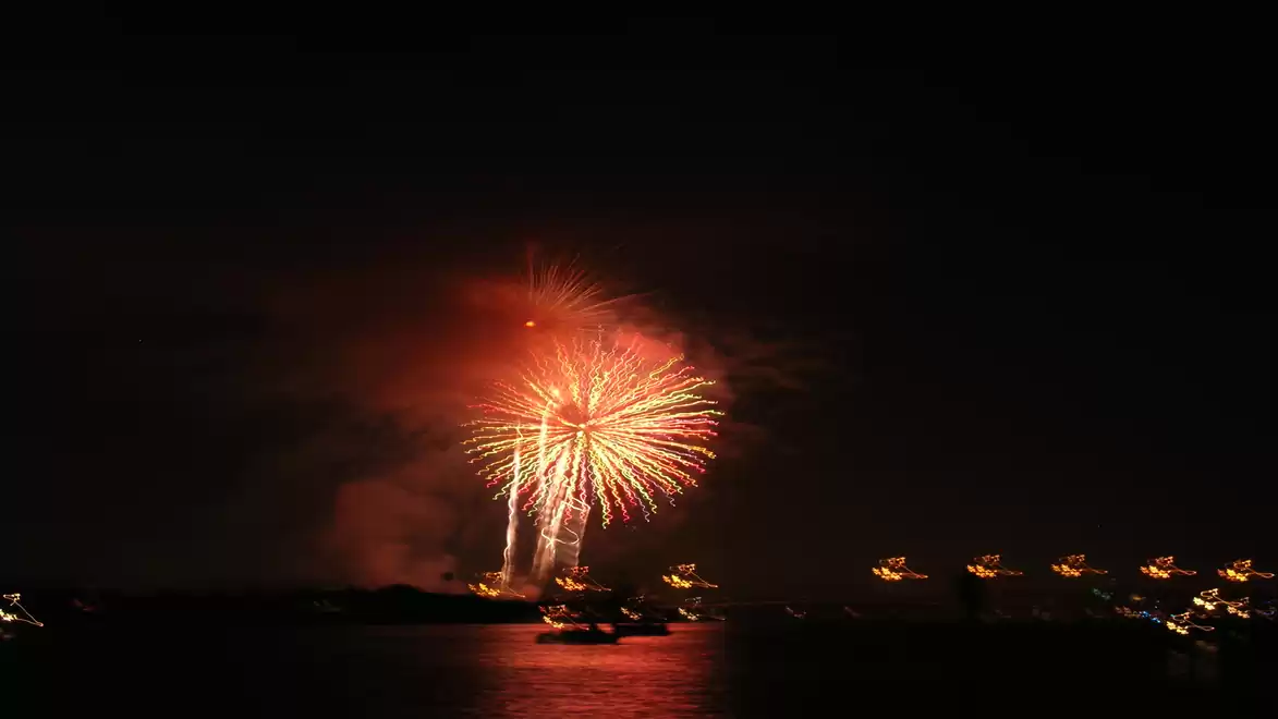 An impressive fireworks display illuminates the night sky over a dark body of water, with lights from boats or buildings visible in the distance.