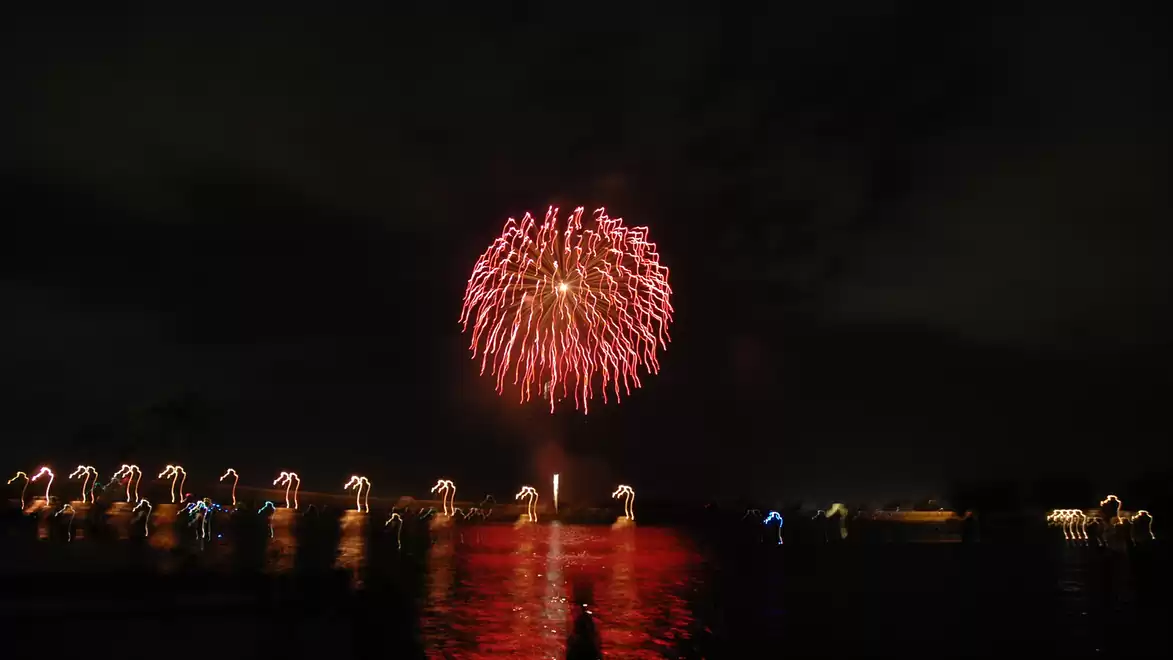 A red firework illuminates the night sky over a body of water with light reflections and silhouetted landscape.