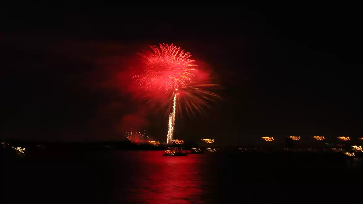 A red firework brightly lights up over a body of water, with lights visible along the shore.