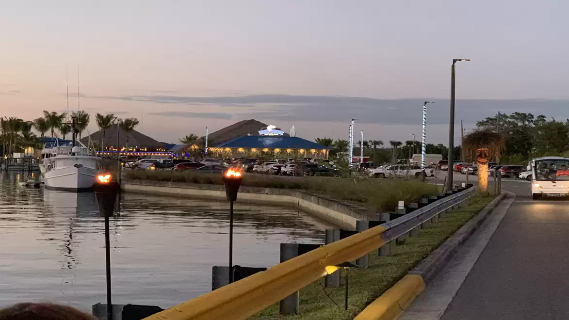 Evening scene at a marina with torches, illuminated buildings, cars in a parking lot, and a boat in the water.