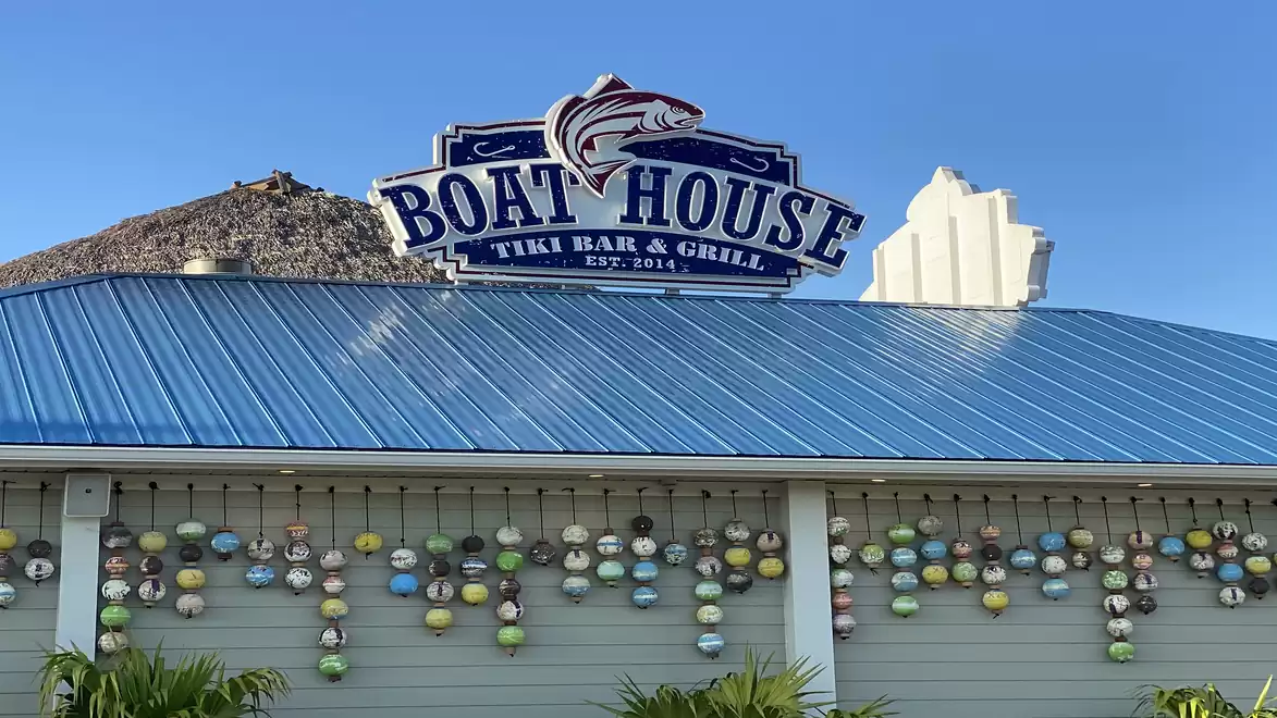 A building with a blue roof featuring a sign that reads 'Boat House Tiki Bar & Grill'. Colorful maritime decorations hang on the facade.