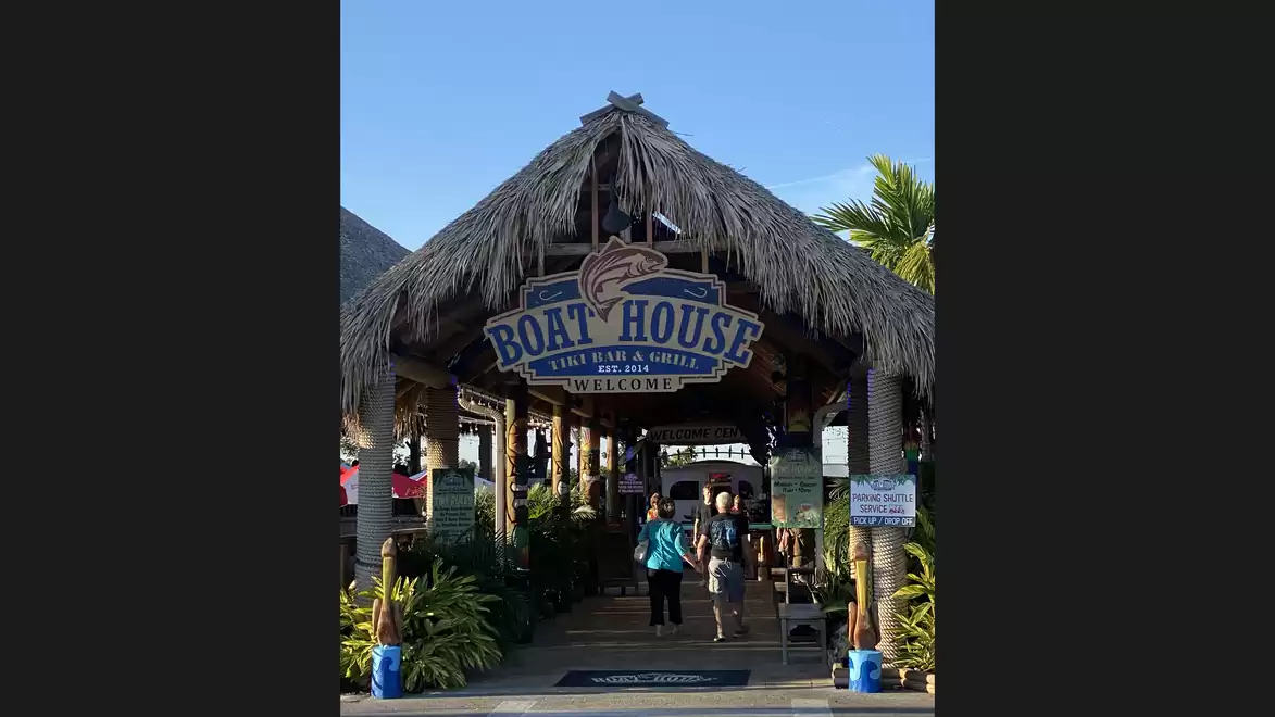 Entrance to a restaurant called 'Boat House', covered with a thatched roof, surrounded by tropical plants and palm trees.