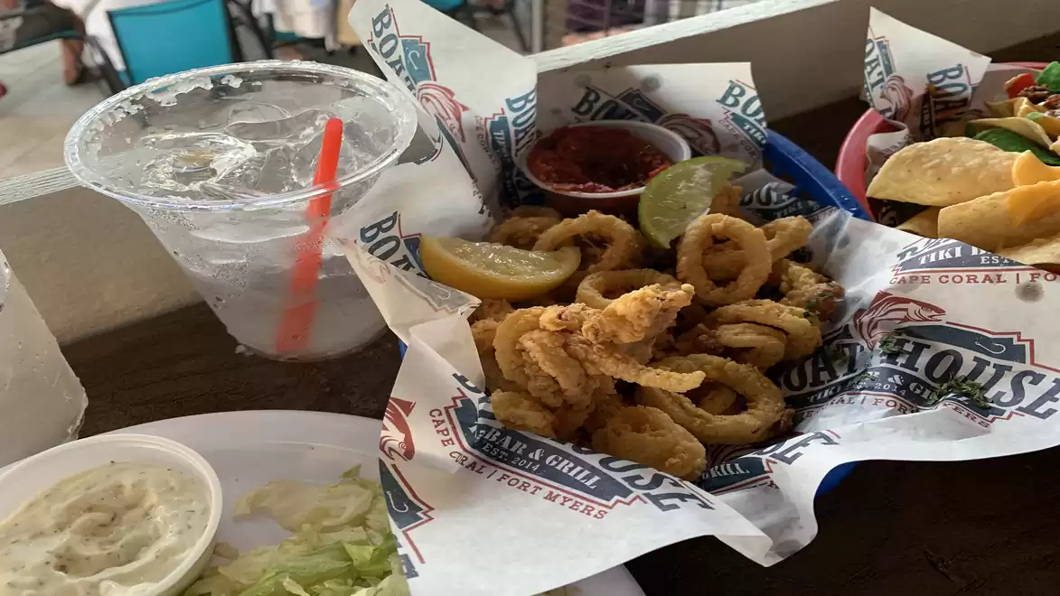 A plate of fried calamari with lemon slices and various sauces at a restaurant, next to a drink with ice cubes and a red straw.