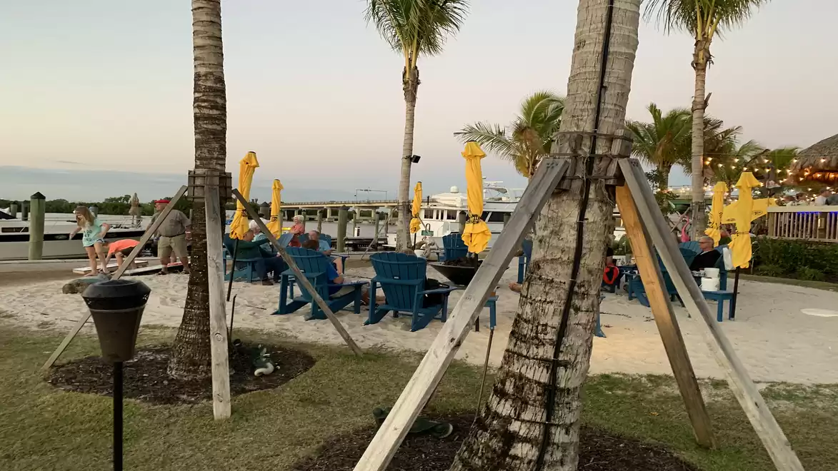 A relaxed outdoor area with palm trees and blue lounge chairs surrounded by yellow umbrellas at sunset on a beach or lake.