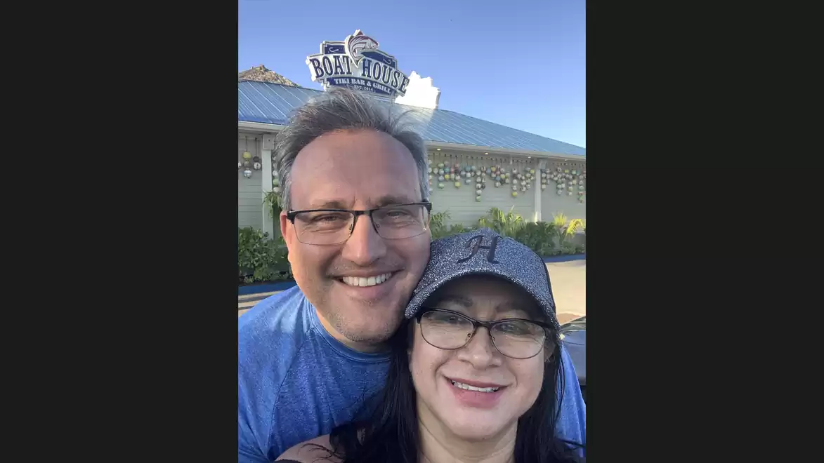A smiling couple stands in front of the Boathouse restaurant with a sunny blue sky in the background.