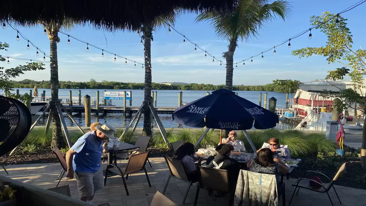 Scene at the waterfront with people dining under palm trees and string lights in an outdoor restaurant, with a calm lake in the background.
