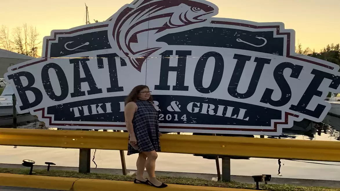 Woman standing in front of a large Boat House Tiki Bar & Grill sign at sunset.