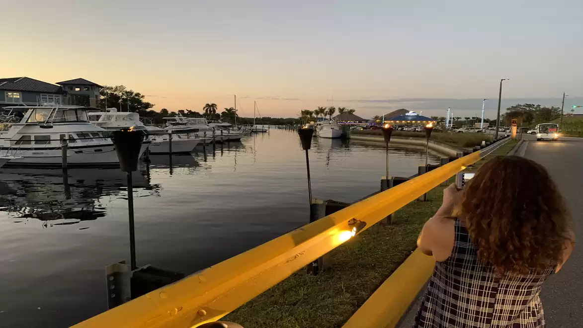 A woman photographs a marina at sunset. Several boats are moored in the water while the sky glows in soft orange and blue tones.