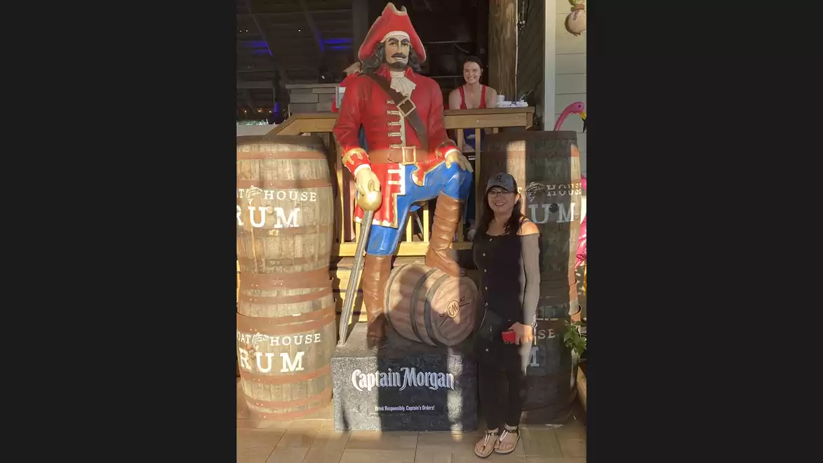 Woman posing next to a large statue of a pirate captain in a red uniform with rum barrels in the background.