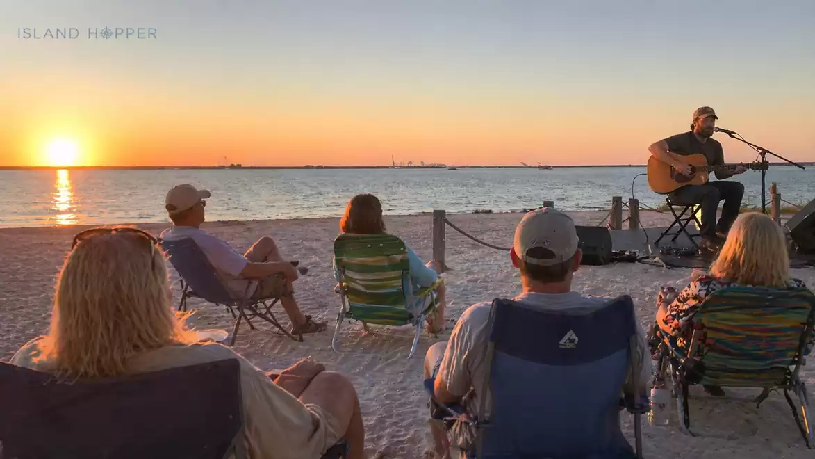 Group of people sitting on beach chairs listening to a guitarist during sunset.