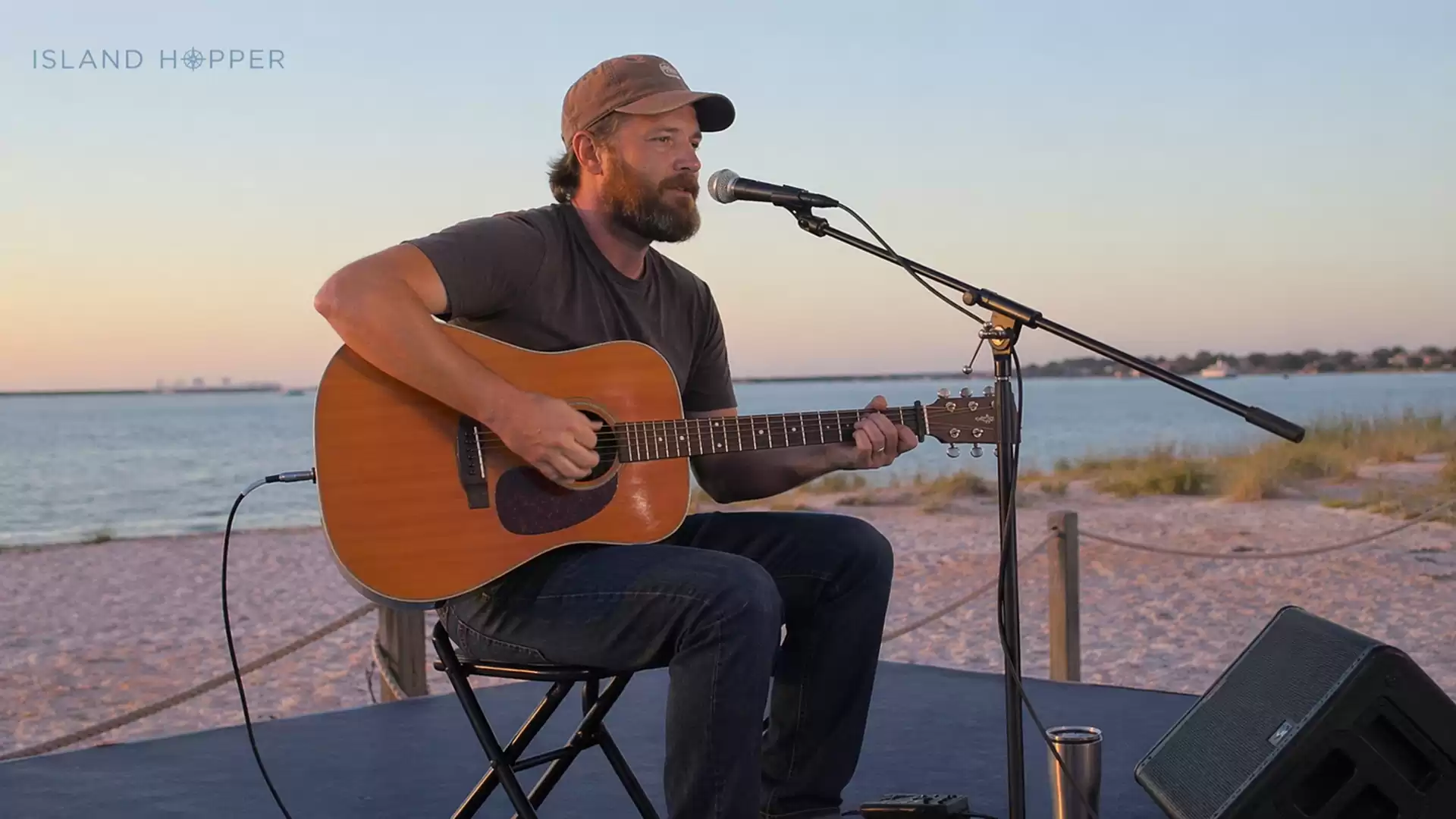 A musician sits on a stage at the beach playing guitar during sunset.
