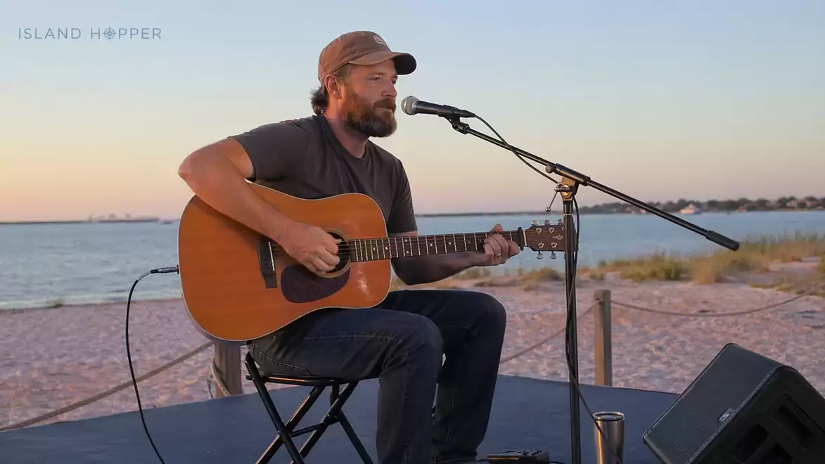 A musician sits on a stage at the beach playing guitar during sunset.