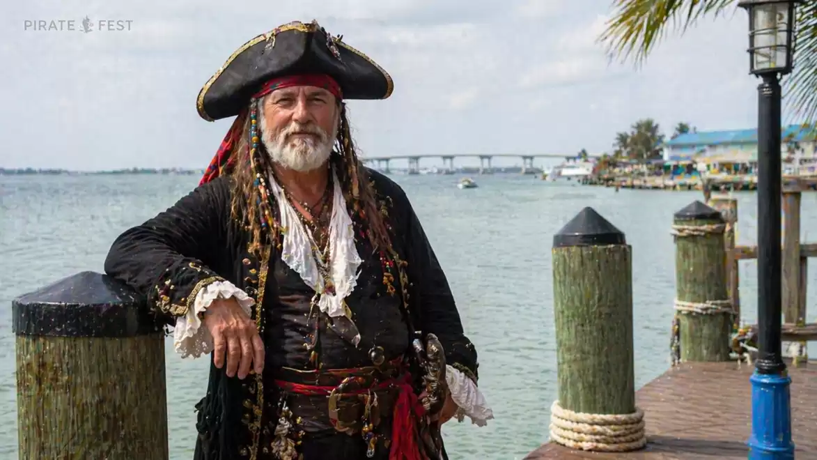 A man in a pirate costume stands at the harbor in front of a maritime backdrop with water and a pier.