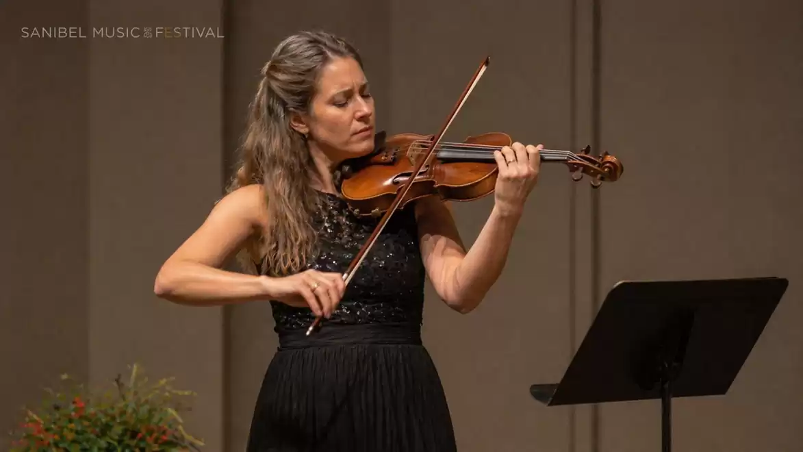 A woman plays the violin intently on stage, dressed in an elegant black dress, during the Sanibel Music Festival.