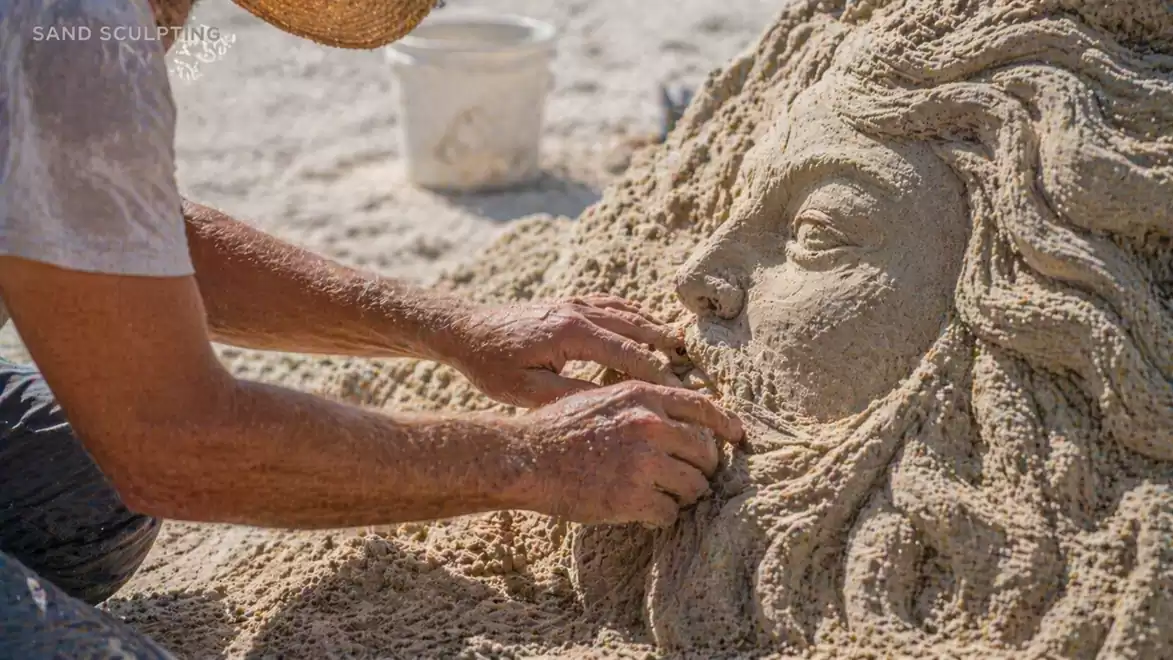 An artist creates an artistic sand face with detailed facial features and hair, with a white bucket in the background.