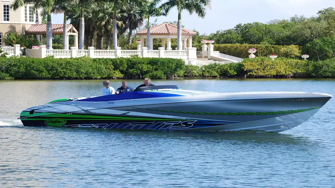 A blue-green motorboat moves on calm water in front of a villa with palm trees in the background.