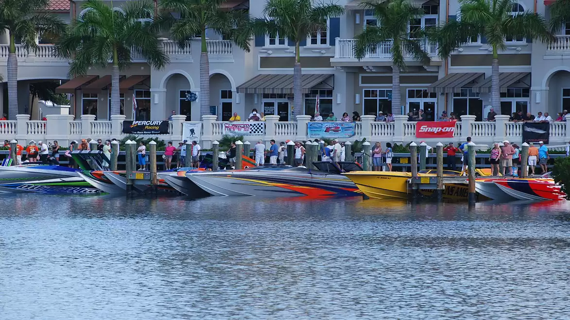 Several colorful speedboats are docked at the pier, with palm trees and buildings in the background. A crowd of people is watching the scene.