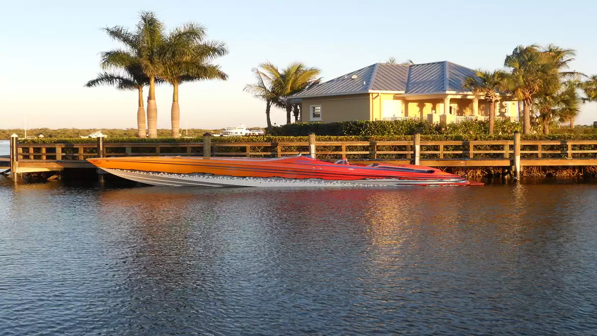 A fast motorboat is moored at a dock, with palm trees and a house in the background.