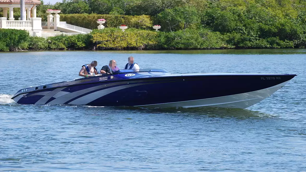 A fast motorboat with four people on board is cruising on a river surrounded by green trees.