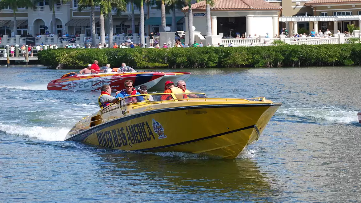 Two fast speedboats, one yellow and one red, cruising on a waterway with lush green trees and a large building in the background.