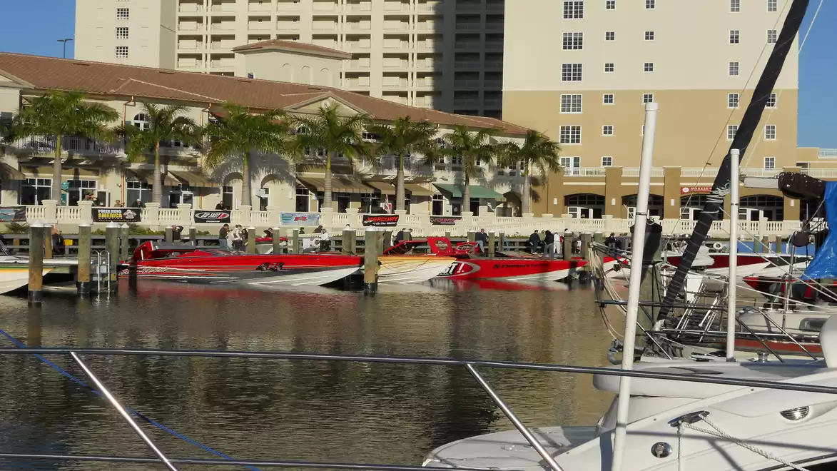 A harbor view with several luxury motor yachts and tall buildings in the background, including palm trees and a clear reflection on the water.