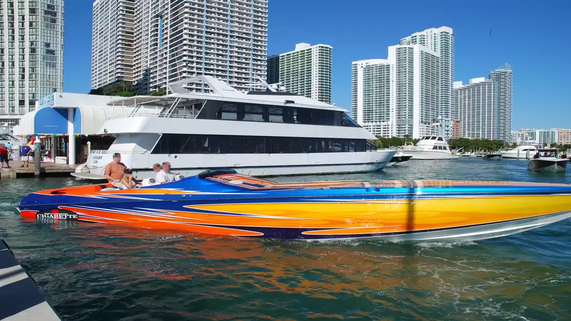 Two boats on the water in front of a skyline of modern skyscrapers. One boat is a large, luxurious yacht, while the other is a colorful speedboat.