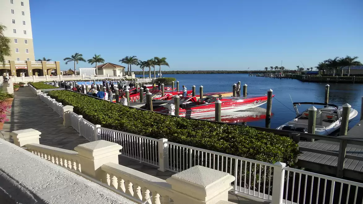 View of a marina with several red boats docked, surrounded by palm trees and a light-colored building in the background.