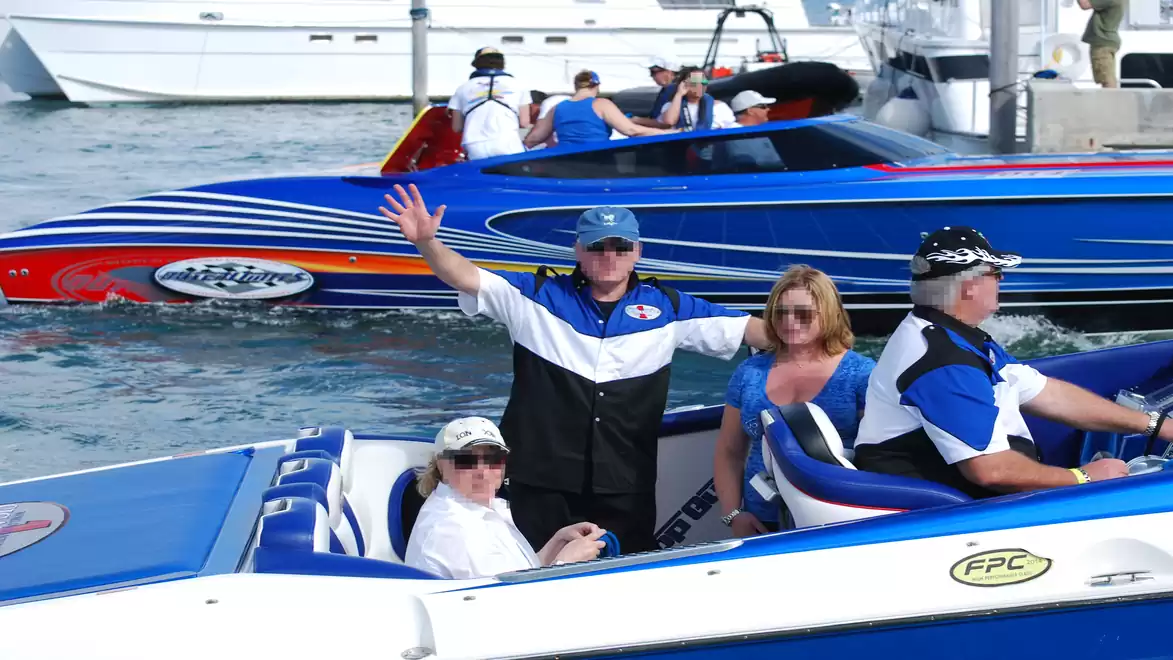 Several people are on a blue speedboat in the water, some sitting and others standing. More boats are visible in the background.