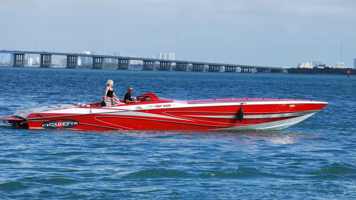 Red motorboat with two people cruising on blue water, with a bridge in the background.