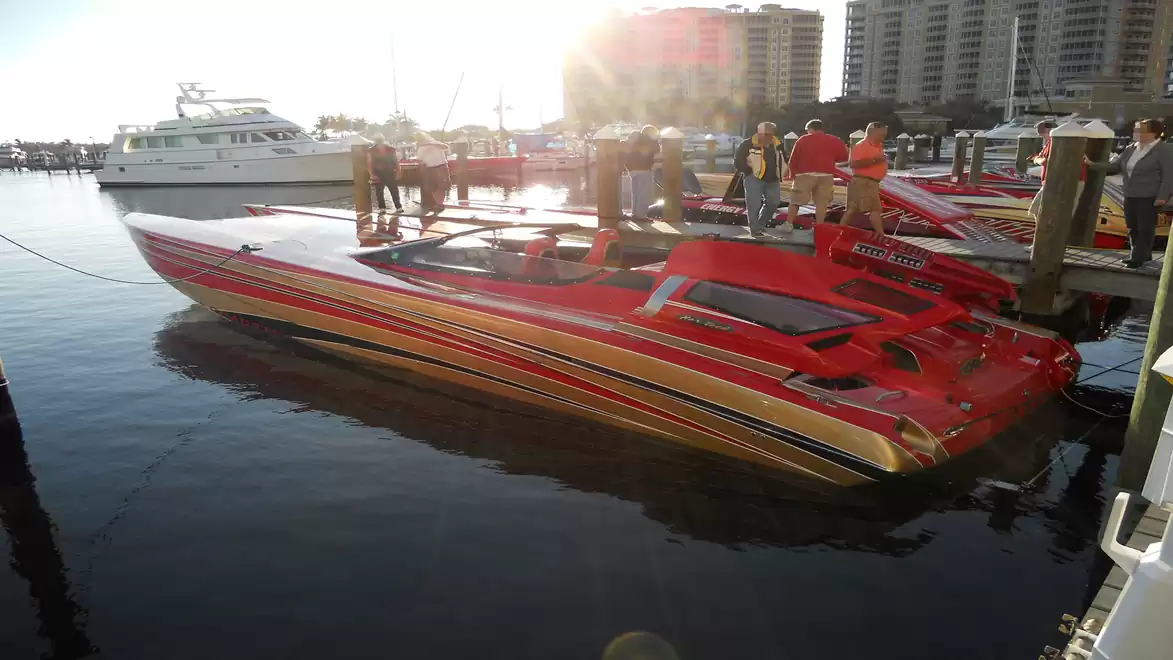 A red speedboat is moored at a dock. Other boats and buildings are visible in the background. Several people are standing on the dock next to the boat.