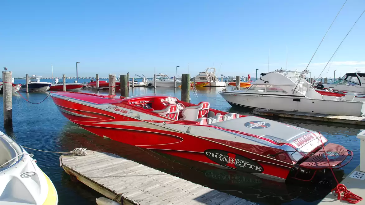 A red speedboat docked at a port on a sunny day next to several other boats. The water is calm and the sky is clear.