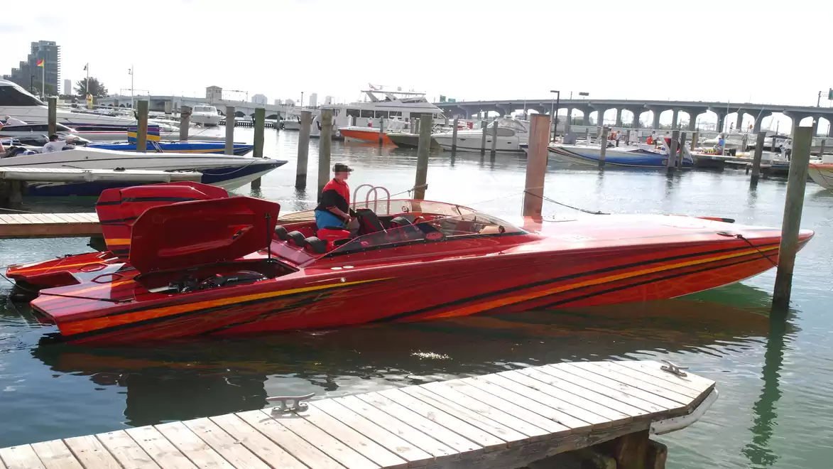 A red speedboat with distinctive orange accents is docked in a harbor. In the background, several boats and a bridge are visible.