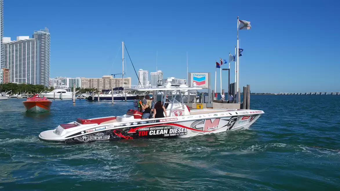 A speedboat with the lettering 'Clean Diesel' is moving on a blue water body near a modern cityscape with skyscrapers and a harbor.