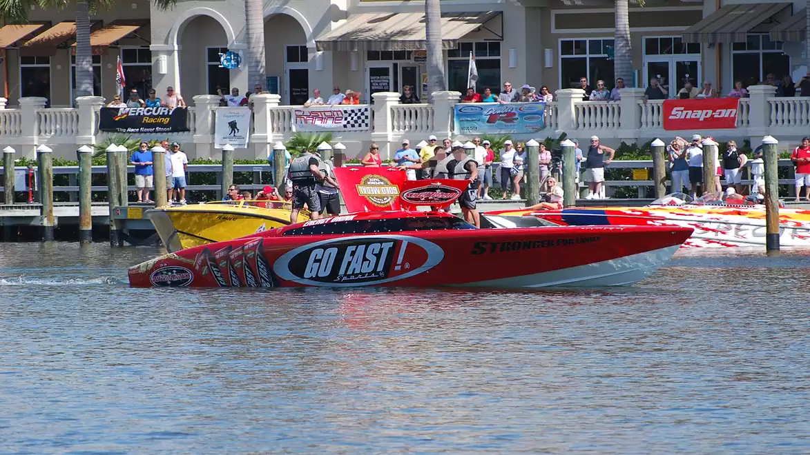 A red speedboat with the words 'Go Fast' floats in front of a dock full of people watching a boat event.