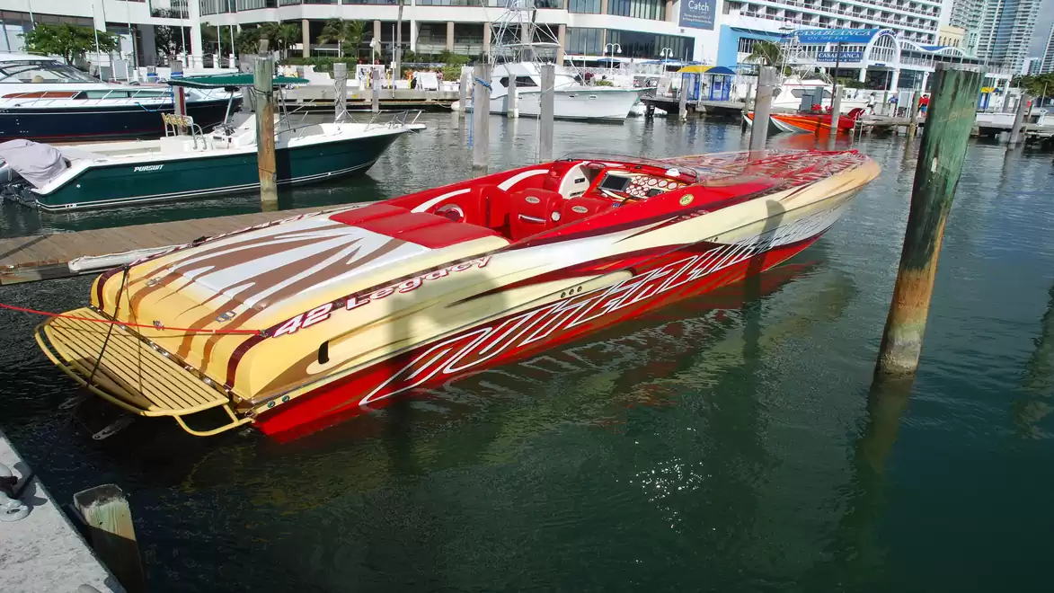 A striking speedboat in red and yellow is docked at a modern marina. Other boats and buildings are visible in the background.