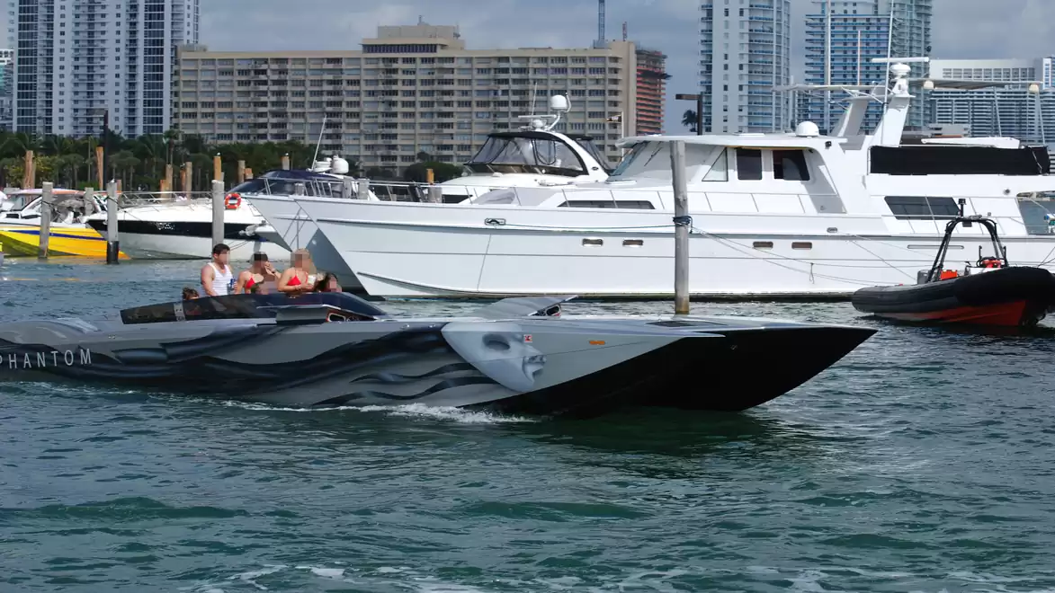 Speedboat on water with several people on board, larger yachts and tall buildings in the background.