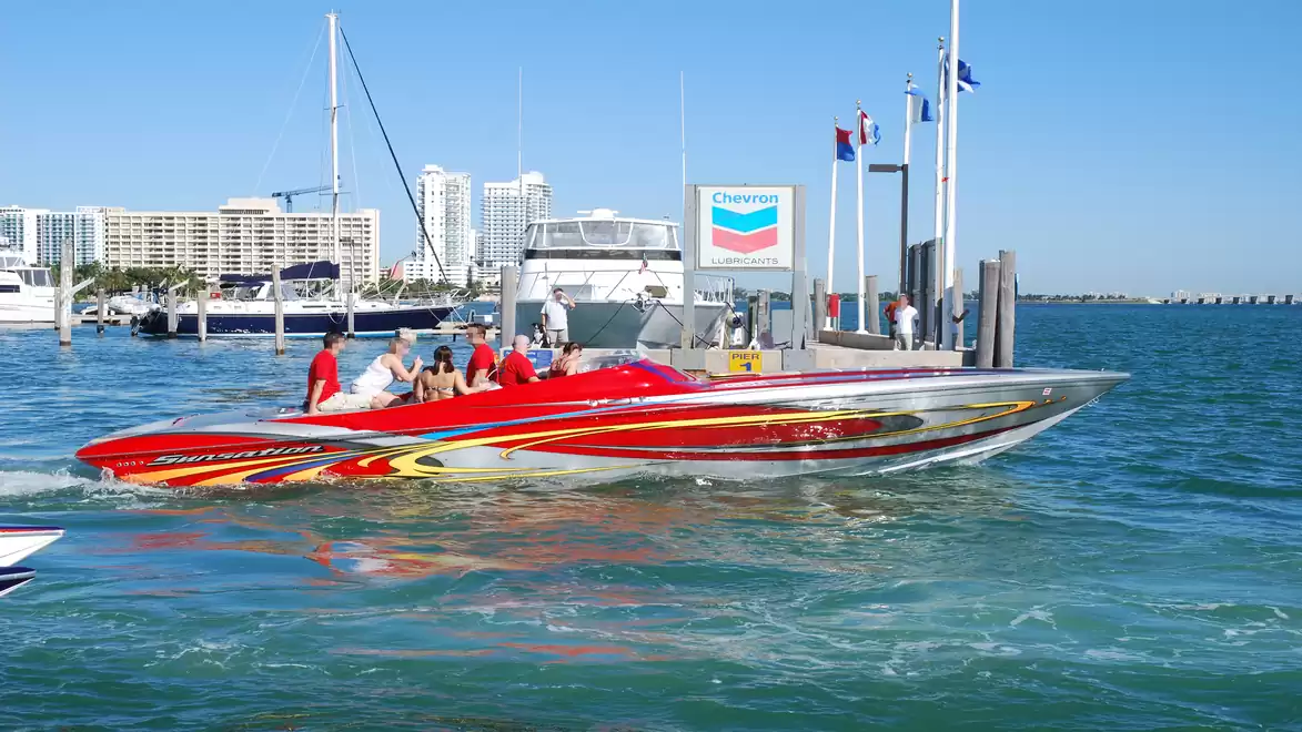 Red speedboat with several people on a body of water near a coastal landscape with marina.