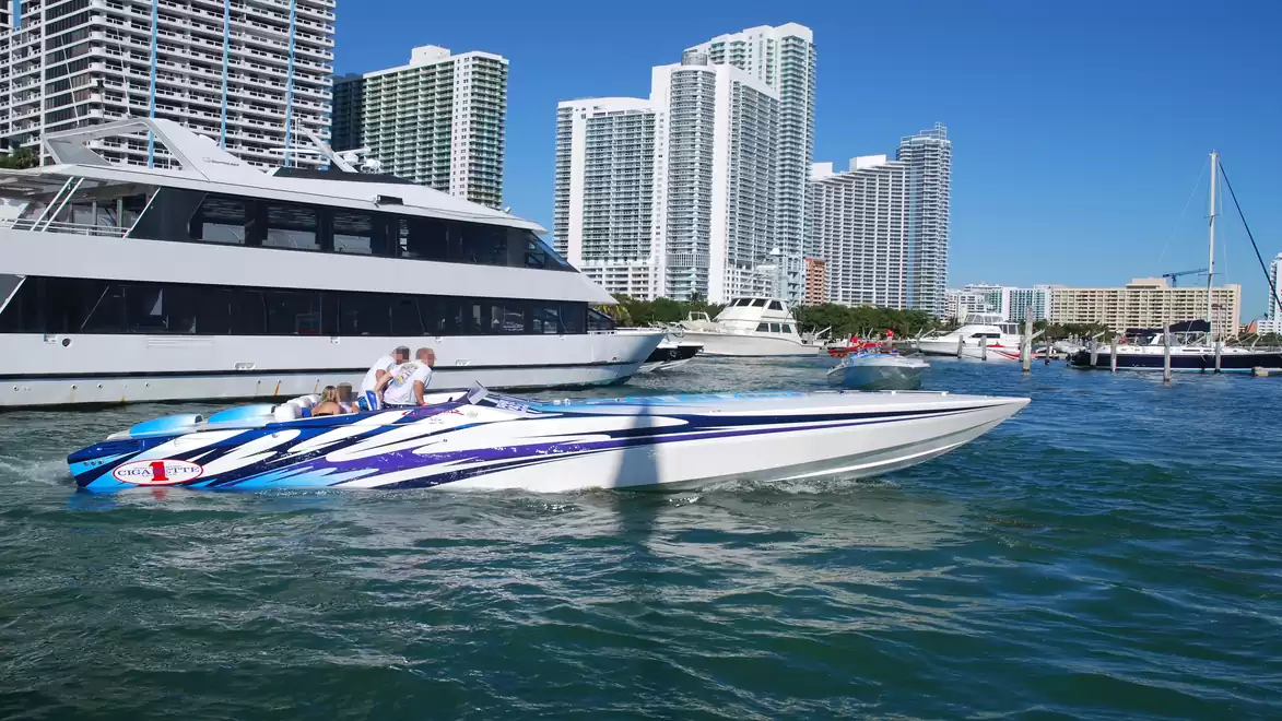 A modern speedboat with blue stripes is sailing in a harbor with tall buildings in the background, while a large yacht is beside it.