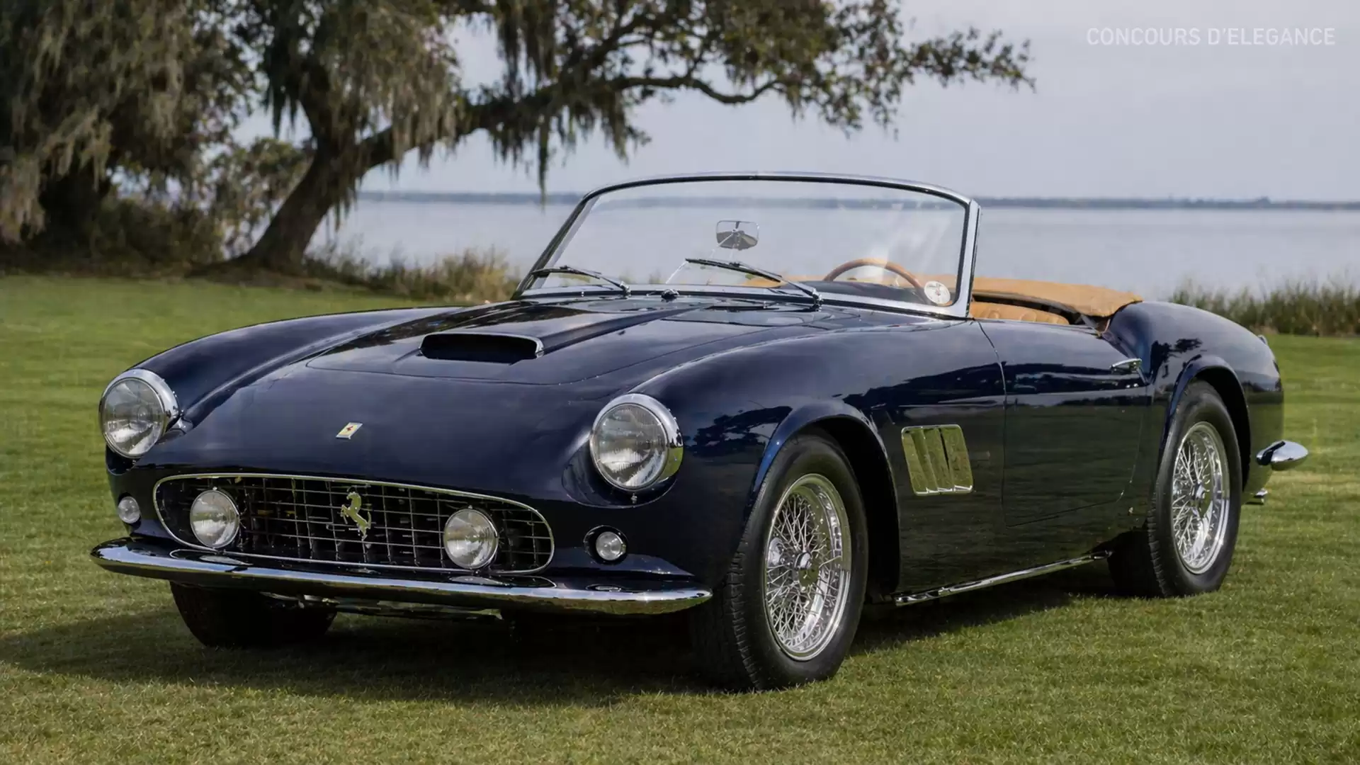 A classic blue sports car is parked on a green lawn with a lake and trees in the background.
