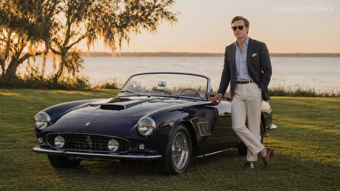 Elegant man in a suit casually stands next to a classic sports car on a green lawn.